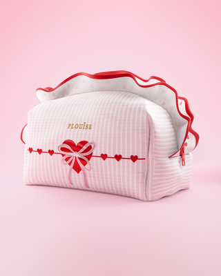 A pink and white striped P.Louise makeup and skincare bag with frills and love heart design on it against a pink background
