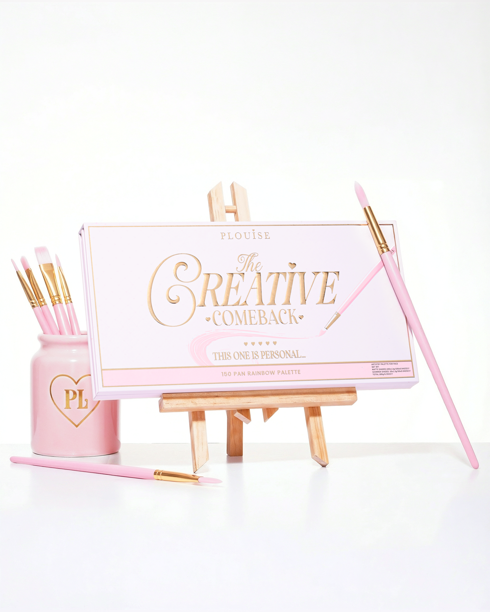 Une palette de fards à paupières arc-en-ciel P.Louise Creative Comeback XXXL rose fermée placée sur un chevalet d'art entouré de pinceaux de maquillage