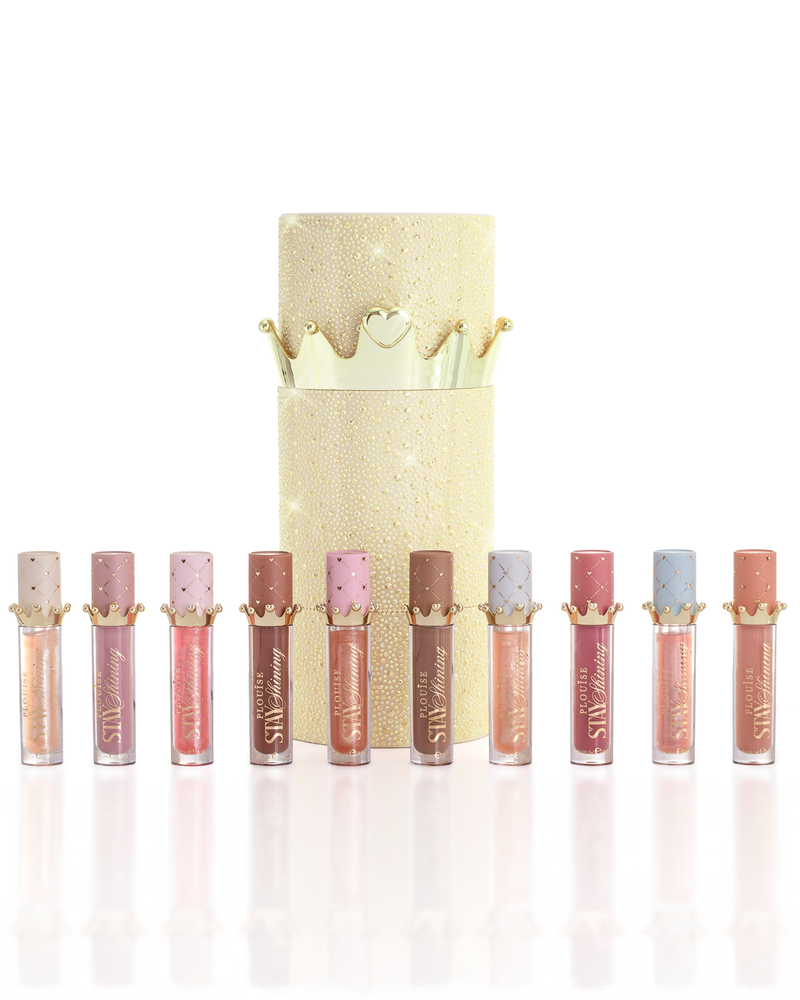 Un ensemble de dix produits d'huile pour les lèvres P.Louise Christmas Stay Shiny disposés en rangée devant une boîte d'huile à lèvres cylindrique dorée pailletée avec un couvercle en forme de couronne dorée, sur un fond blanc propre.