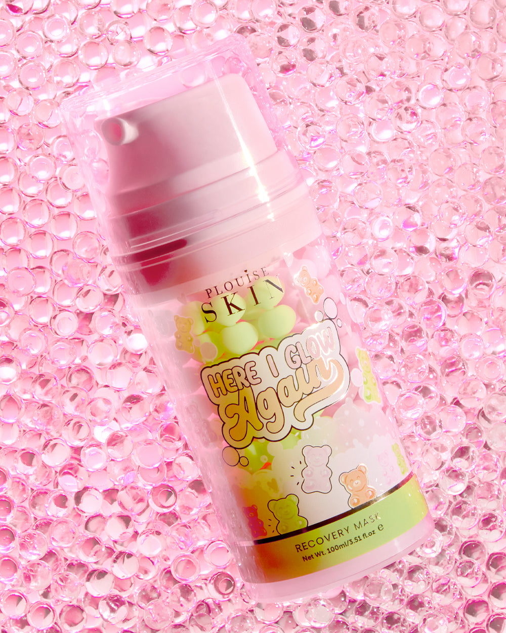 Une bouteille de masque de récupération P.Louise Here I Glow Again au parfum bonbons ours avec une étiquette rose et verte, avec des illustrations de bonbons ours, placées sur un fond rose pailleté.