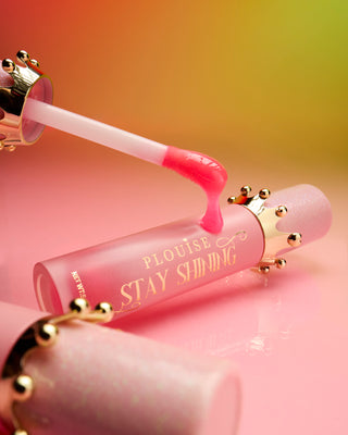 A close-up image of a P.Louise Stay Shining Crown Lip Oil bottle with a gold-coloured crown around the cap and a pink applicator wand, showing the product's texture and subtle pink colour.