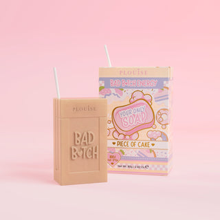 A beige bar of soap with 'Bad Bitch' engraved on it, accompanied by a colourful juice box package with 'Piece of Cake' labelled on it, presented on a pink background.